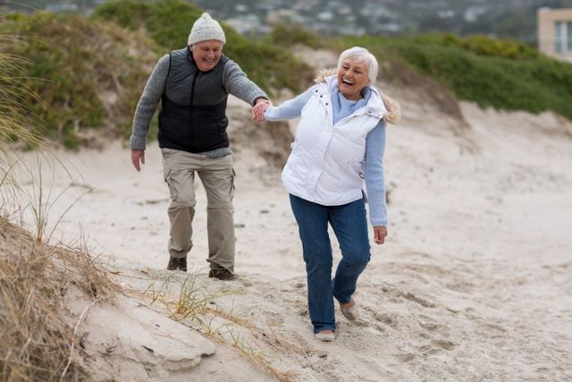 Senior couple walking on beach, holding hands and smiling. Perfect for use in advertisements for retirement communities, health and wellness campaigns, travel brochures, and lifestyle blogs focusing on active aging and senior living.