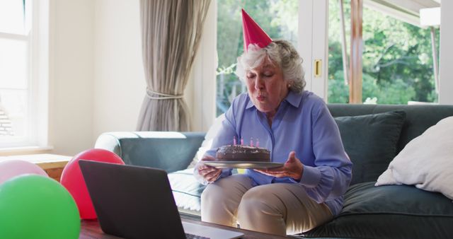 Caucasian senior woman in party hat blowing candles on birthday cake while having a image call on laptop at home. social distancing during coronavirus quarantine lockdown.