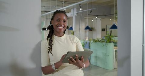 Smiling professional woman holding tablet and leaning in modern coworking office with greenery