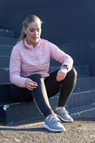 Young woman resting on urban steps after workout wearing pink jacket and wireless earbuds