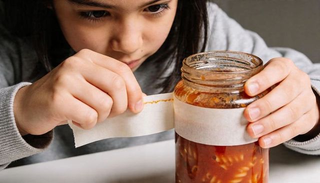 Curious girl peeling label from pasta sauce jar with messy tomato hands, intimate closeup