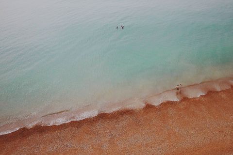 Aerial turquoise sea meeting pebbled red beach with people wading and paddling
