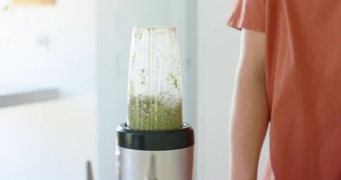 Teenage male in orange tee making green smoothie with personal blender in sunlit modern kitchen