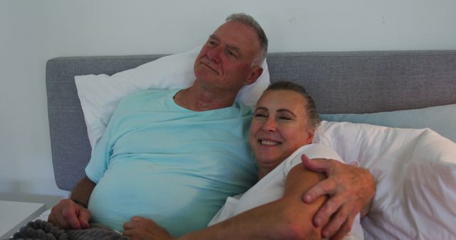 Elderly couple relaxing in bed, smiling and embracing. This image can be used for advertisements, healthcare services targeting senior citizens, retirement planning promotions, and lifestyle blogs discussing intimacy and relationships among aging populations.
