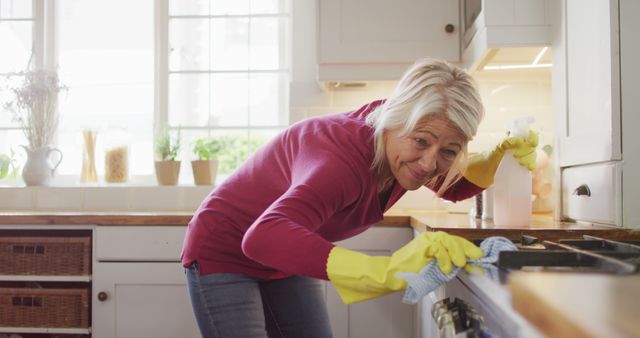 Senior woman smiling while cleaning kitchen stove. She is wearing yellow gloves and appears to be enjoying the task. Ideal for articles or advertisements related to housework, home cleaning products, senior lifestyle, domestic chores, and hygiene.
