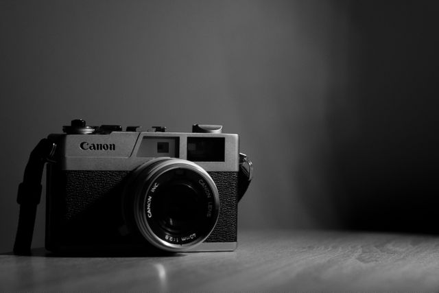 Detailed image of a vintage Canon film camera placed on a wooden surface with a dramatic monochrome background. Ideal for content related to photography, technology, vintage items, and retro aesthetics. Great for use in articles, blogs, and marketing materials celebrating the history and evolution of cameras and photography.
