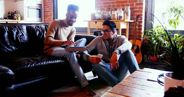 Two young friends together in a stylish modern apartment, sitting in a cozy living room with exposed brick walls and plants. One is tapping on a tablet while the other engages with a smartphone, capturing technology and social interaction. Ideal for concepts of leisure, modern lifestyle, technology usage, friendship, home decor, and relaxation.