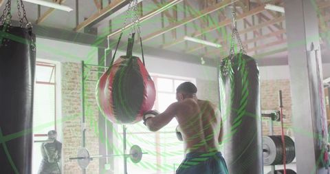 Shirtless boxer training and punching red heavy bag in sunlit industrial gym with brick walls