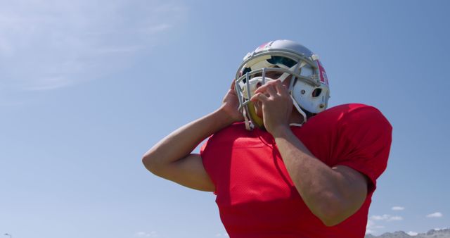 Football player in red jersey adjusting helmet on a sunny day. Ideal for depicting sports themes, athletic preparation, and outdoor activities. Suitable for use in sports marketing, fitness campaigns, and youth sports promotions.