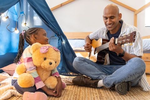 Asian father playing guitar with african american daughter in cozy bedroom tent