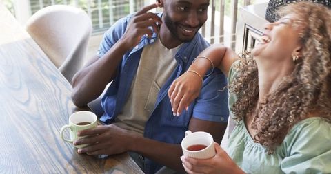 Diverse couple sharing morning coffee and laughing at wooden table in cozy sunlit home