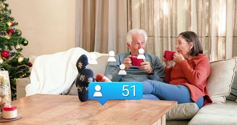 Senior couple sharing cozy holiday coffee on sofa, sipping red mugs with 51 notifications