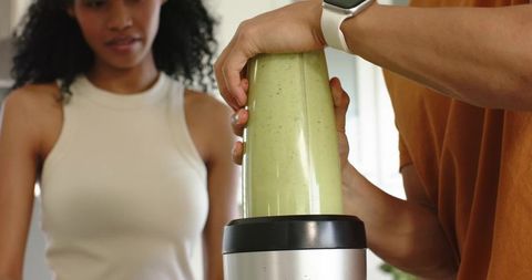 Diverse couple making green smoothie with portable blender on sunlit kitchen countertop