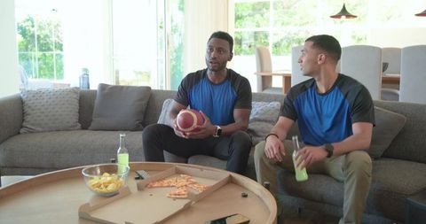 Diverse friends watching football at home wearing matching shirts holding ball and drinks