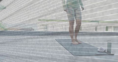 Woman practicing rooftop yoga standing on mat with towel and water bottle on urban terrace