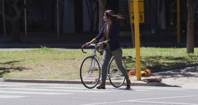 Woman in casual clothing walking with a bicycle on a city street crosswalk. She is engaged in urban commuting, emphasizing safety by walking through the pedestrian lane. Ideal for use in content related to commuting, urban living, transportation, safety, and eco-friendly travel options.