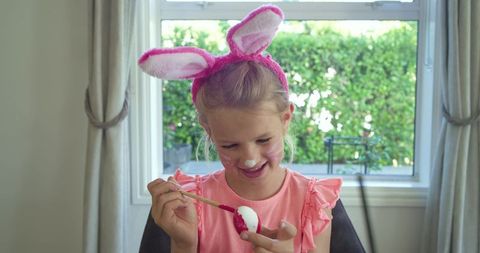 Little girl painting easter egg wearing pink bunny ears by sunlit window