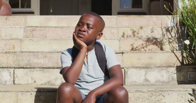 Image shows a young boy sitting on stone steps, resting his chin on his hand and appearing thoughtful. He wears a light blue shirt and dark shorts, with a backpack on his shoulders, suggesting he is a student. The scene is bathed in bright sunlight, with part of a house and greenery visible in the background. This can be used to depict themes of reflection, childhood, school days, waiting, or calm moments.