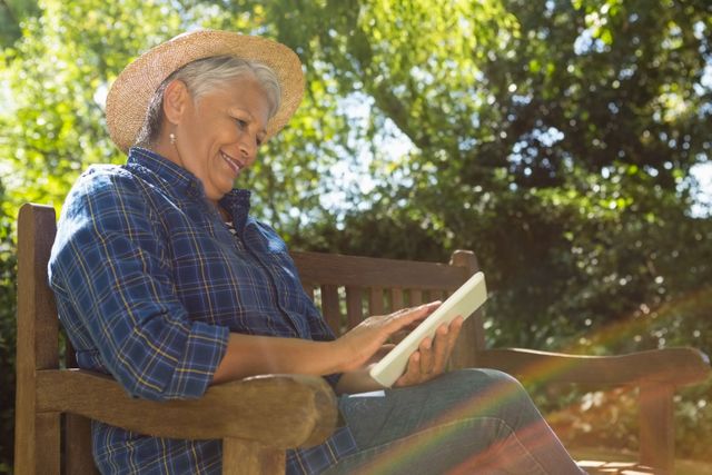 Senior woman enjoying a digital book on a tablet while sitting on a wooden bench in a garden. Ideal for themes related to senior lifestyle, technology use among elderly, relaxation, and outdoor activities. Perfect for articles, advertisements, and blogs focusing on retirement, leisure, and digital literacy for seniors.