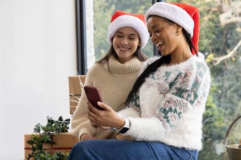 Multiracial women wearing santa hats sharing phone and smiling during cozy holiday moment