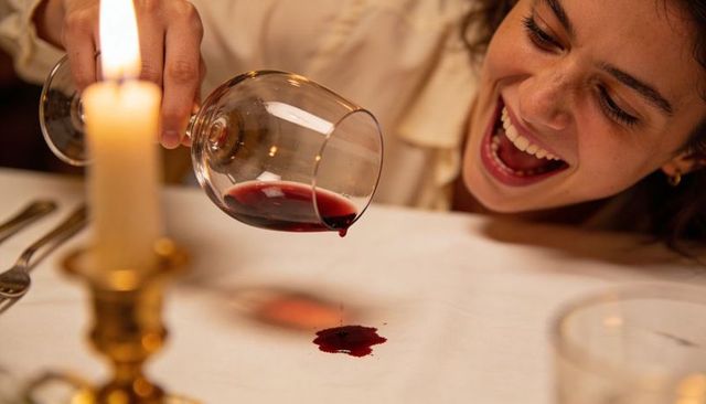 Candlelit dinner mishap: laughing woman tilting wine glass, red wine spilling on linen