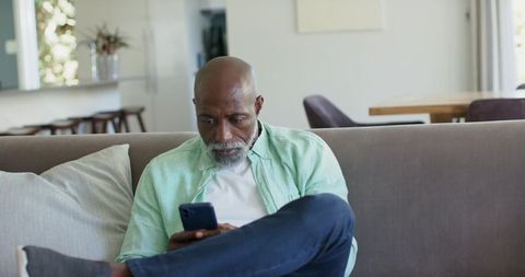African american senior man relaxing on sofa using smartphone in modern living room