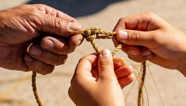 Weathered hands teaching child to tie braided rope knot on sunlit sandy ground