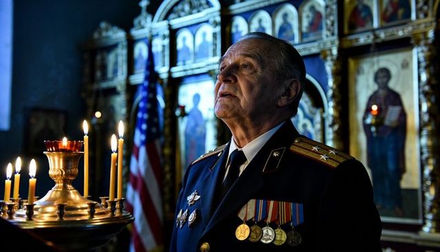 Elderly veteran standing in orthodox church wearing medals and uniform, gazing upward