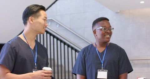 Multicultural male nurses chatting on hospital staircase holding coffee cups and badges