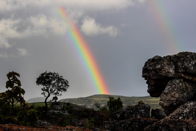 Rainbow arching over a rugged rocky landscape, complemented by a solitary tree and mountainous terrain in the distance. Perfect for nature-themed projects, travel brochures, or inspirational visuals.