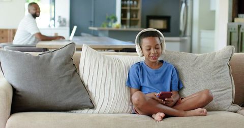 African American boy using smartphone with headphones while father works on laptop