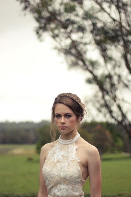 Young bride standing in a garden, wearing an elegant lace wedding dress with a high neckline and an intricate hairstyle. She shows a serene and sophisticated look, perfect for wedding-related marketing, wedding magazines, bridal fashion blogs, and engagement announcements. Captures outdoor wedding themes and bridal elegance.