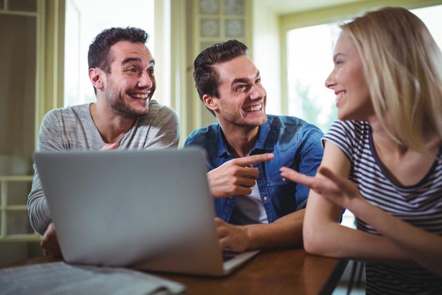 Three friends smiling and enjoying a casual conversation while using a laptop at a café. Ideal for themes of friendship, social interactions, technology use in social settings, and modern lifestyles.