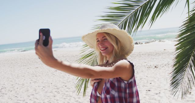 Young woman wearing straw hat smiling and taking selfie on a sunny beach with palm leaves and ocean in the background. Ideal for portraying summer vacation, beach holidays, travel, social media, and outdoor activities.