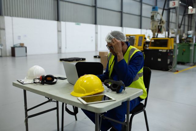 Side view of a focused biracial male worker, busy working in a factory warehouse, wearing a hair net, bue overalls and a high visibility vest, sitting and using laptop computer, with his hard hat and a tablet on the table