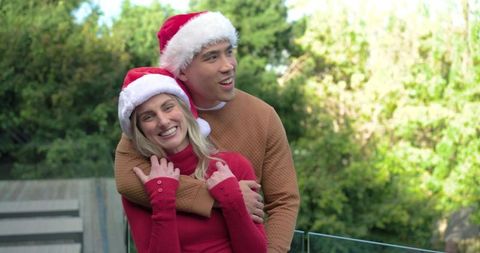 Young couple wearing santa hats hugging on glass balcony with holiday sunlight and greenery