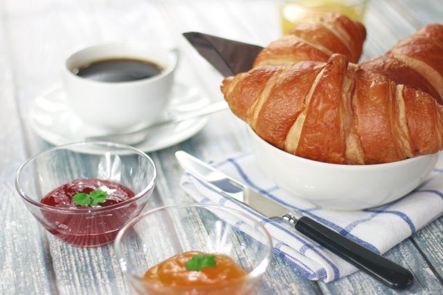 Freshly baked croissants in a bowl alongside a cup of black coffee, butter knife, and small bowls of jam on a grey wooden table. Perfect for illustrating breakfast scenes, morning meals, bakery promotions, or dining setups.