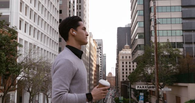 Picture of a young professional enjoying coffee while standing in an urban landscape. Dressed casually, he projects a modern urban lifestyle. Useful for themes related to business, city living, young professionals, and casual modern lifestyles. Ideal for stories and campaigns featuring an urban environment or business lifestyle.