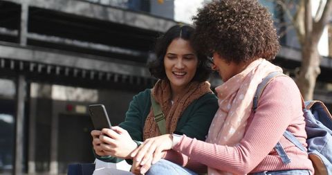 Two friends sharing phone and laughing on urban bench with backpacks and scarves