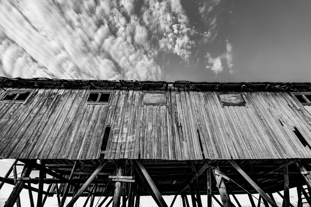 Old wooden house elevated on stilts, captured in black and white with clouds in the sky. The image has a high contrast, emphasizing the texture of the wood and the drama of the sky. Suitable for themes about historical structures, rural life, decay, architecture, and photography art.