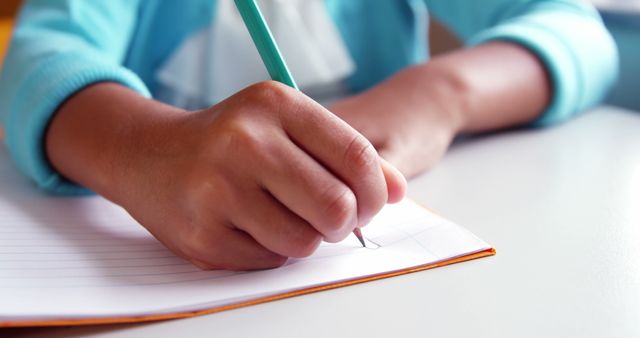 Close-up of child's hand writing in notebook with pencil on desk. Useful for educational materials, school promotions, learning resources, and child development content.