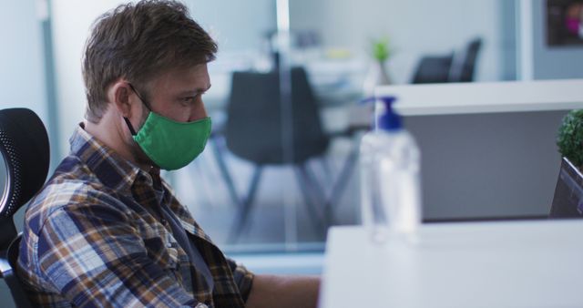 Man in an office setting wearing a green face mask working on a computer. A hand sanitizer bottle is visible on the desk, highlighting health and safety measures during the COVID-19 pandemic. This image can be used to illustrate workplace safety, pandemic precautions, and remote or office work environments.