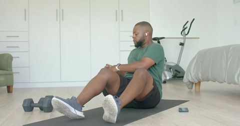 Man checking smartwatch during home workout on mat with dumbbells and exercise bike