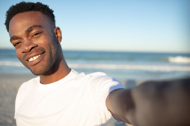 Young man enjoying a sunny day at the beach, capturing a selfie with the ocean in the background. Ideal for use in travel blogs, vacation advertisements, lifestyle articles, and social media promotions highlighting leisure and relaxation.