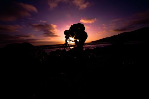 Silhouette photographer capturing vibrant sunset over ocean on rocky shore with starburst