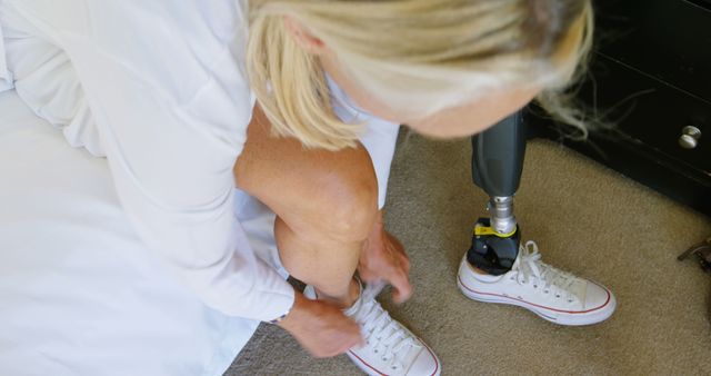 Woman with prosthetic leg tying her shoes in bedroom illustrates determination and independence. Suitable for use in healthcare, disability awareness, and motivational content. Neutral and relatable setting for lifestyle and daily routine visuals.