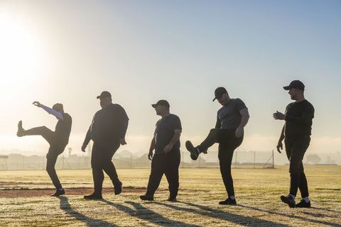 Team warmup at sunrise: men performing synchronized leg kicks on dewy sports field