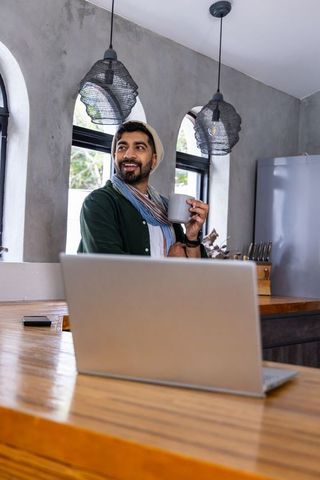 Smiling indian man working from home in modern kitchen holding coffee mug by laptop