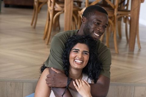 Black man embracing south asian woman while smiling on wooden step at home, cozy romance