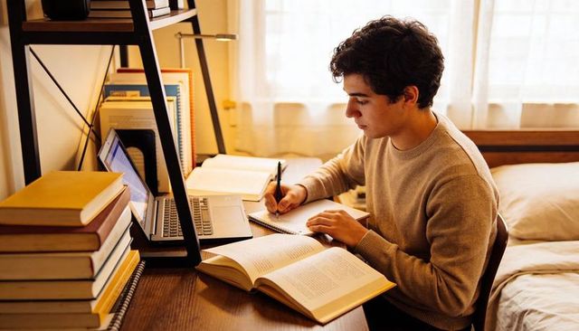 College student studying with laptop and textbooks at cozy bedroom desk in warm natural light